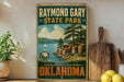 A vintage-style sign for Raymond Gary State Park in Oklahoma, featuring a scenic landscape with a lake, mountains, and a sailboat.