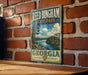 A vintage-style metal sign for Reed Bingham State Park in Georgia, featuring a scenic landscape with a lake and mountains.
