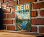 A vintage-style sign for Reid State Park in Maine, featuring a scenic landscape with pine trees, a lake, and mountains in the background.