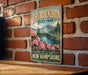 A vintage-style sign for Rhododendron State Park in New Hampshire, featuring a scenic landscape with mountains, a river, and pink flowers.