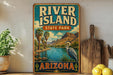 A vintage-style sign for River Island State Park in Arizona, featuring a scenic landscape with palm trees, mountains, and a heron.