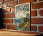 A vintage-style metal sign for Robert Louis Stevenson's State Park in California, featuring a scenic landscape with mountains, a river, and a path leading to the park.