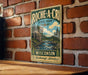a vintage-style metal sign with a scenic illustration of a river and forest, mounted on a brick wall.