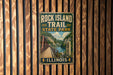 A vintage-style metal sign for Rock Island Trail State Park in Illinois, featuring a scenic illustration of a bridge and a river.