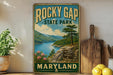 A vintage-style sign for Rocky Gap State Park in Maryland, featuring a scenic mountain landscape with a lake and pine trees.