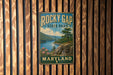 A vintage-style metal sign for Rocky Gap State Park in Maryland, featuring a scenic landscape with mountains, a lake, and a pine tree.
