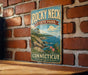 A vintage-style sign for Rocky Neck State Park in Connecticut, featuring a scenic coastal landscape with a rocky shoreline, a sailboat on the water, and a cloudy sky.