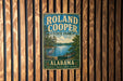 A vintage-style metal sign for Roland Cooper State Park in Alabama, featuring a scenic illustration of a lake, trees, and a bird.