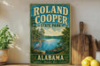 A vintage-style sign for Roland Cooper State Park in Alabama, featuring a scenic illustration of a lake, a crane, and a canoe.