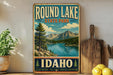 A vintage-style sign for Round Lake State Park in Idaho, featuring a scenic mountain landscape with a lake and pine trees.