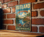 A vintage metal sign for Rutland State Park in Massachusetts, featuring a scenic landscape with pine trees, a lake, and mountains.