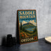 A vintage metal sign for Saddle Mountain State Natural Area in Oregon, featuring a scenic mountain landscape with pine trees and a lake.