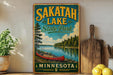 A vintage-style sign for Sakatah Lake State Park in Minnesota, featuring a red canoe on a lake surrounded by pine trees and a cloudy sky.