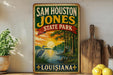A vintage-style metal sign for Sam Houston Jones State Park in Louisiana, featuring a scenic illustration of a sunset over a body of water with a crane in the foreground.