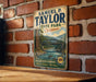 A vintage-style metal sign for Samuel P. Taylor State Park in California, featuring a scenic illustration of a river and bridge surrounded by redwood trees.