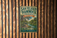 A vintage-style metal sign for San Felasco Hammock Preserve, featuring a tropical landscape with palm trees and a river.