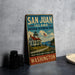 A vintage-style sign for San Juan Island National Historical Park, featuring a lighthouse and a house, is displayed on a gray countertop next to some black camera lenses.
