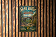 A vintage-style metal sign for Sand Bridge State Park in Pennsylvania, featuring a scenic illustration of the bridge surrounded by lush greenery.
