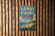 A vintage-style metal sign for Sandy Point State Park in Maryland, featuring a lighthouse and beach scene, is displayed on a wooden wall.