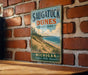 A vintage metal sign for Saugatuck Dunes State Park in Michigan, featuring a scenic beach landscape and the text "Michigan Explore. Relax. Discover."
