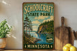 A vintage-style sign for Schoolcraft State Park in Minnesota, featuring a scenic landscape with pine trees, a lake, and mountains.