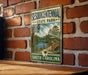 A vintage-style metal sign for Sesquicentennial State Park in South Carolina, featuring a scenic illustration of a bridge, trees, and a lake.