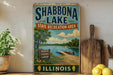A vintage-style sign for Shabbona Lake, Illinois, featuring a scenic image of the lake and surrounding landscape.