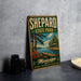 A vintage-style metal sign for Sheppard State Park in Mississippi, featuring a scenic landscape with a dock and trees, is displayed on a gray countertop next to a stack of black camera lenses.
