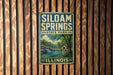 A vintage metal sign for Siloam Springs State Park in Illinois, featuring a scenic landscape with a waterfall and a small cabin.