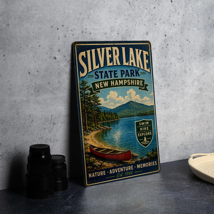A vintage-style metal sign for Silver Lake State Park in New Hampshire, featuring a scenic image of a lake, mountains, and a canoe.