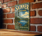 A vintage-style sign for Skinner State Park in Hadley, Massachusetts, featuring a scenic landscape with a lake and mountains.