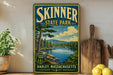A vintage-style sign for Skinner State Park in Hadley, Massachusetts, featuring a scenic landscape with a lake and mountains.