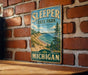 A vintage-style metal sign for Sleeper State Park in Michigan, featuring a scenic landscape with a beach, mountains, and pine trees.