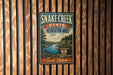 A vintage-style metal sign for Snake Creek State Recreation Area in South Dakota, featuring a scenic landscape with a river, mountains, and pine trees.