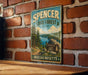 A vintage metal sign for Spencer State Forest in Massachusetts, featuring a scenic landscape with pine trees, a lake, and mountains.