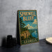 A vintage-style sign for Sprewell Bluff State Park in Georgia, featuring a scenic landscape with a river and cliffs.