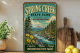 A vintage-style sign for Spring Creek State Park in Washington, featuring a scenic landscape with a river, mountains, and pine trees.