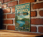 A vintage-style metal sign for Squantz Pond State Park in Connecticut, featuring a scenic image of the pond surrounded by trees and mountains.