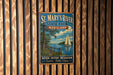 A vintage-style metal sign for St. Mary's River State Park, Maryland, featuring a sailboat on a lake and a wooden fence in the background.