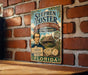 A vintage-style metal sign for Stephen Foster's Folk Culture Center in Florida, featuring a portrait of a man and an illustration of a house.