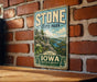 A vintage-style metal sign for Stone State Park in Iowa, featuring a scenic landscape with a river and mountains.