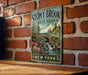 A vintage-style metal sign for Stony Brook State Park in New York, featuring a scenic landscape with a bridge, waterfalls, and trees.