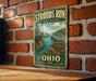 A vintage-style metal sign for Strouds Run State Park in Ohio, featuring a scenic landscape with a river and mountains.