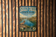 A vintage metal sign for Sully Creek State Park in North Dakota, featuring a scenic illustration of a mountain landscape with a river.