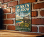 A vintage-style sign for Sunken Meadow State Park in New York, featuring a scenic illustration of the park's landscape with a beach, flowers, and a sign indicating the location.