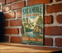 A vintage-style metal sign for Sycamore State Park in Ohio, featuring a scenic landscape with a lake and trees, mounted on a brick wall.