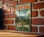 A vintage-style metal sign for Taun sauk mountain in Missouri, featuring a scenic landscape with pine trees and a rocky outcrop.