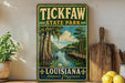 A vintage-style sign for Tick Faw State Park in Louisiana, featuring a scenic illustration of a bayou landscape with a wooden walkway and a bridge.