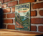 A vintage-style metal sign for Tishomingo State Park in Mississippi, featuring a scenic landscape with a river, mountains, and pine trees.