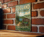A vintage metal sign for Tolland State Forest in Massachusetts, featuring a scenic landscape with pine trees, a lake, and mountains.